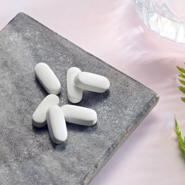Sechs weiße Herbalife Xtra-Cal Tabletten liegen auf einer grauen Steinplatte neben einem Glas Wasser und einem grünen Farnzweig, um die natürliche Zufuhr von Calcium und Vitamin D zu visualisieren.