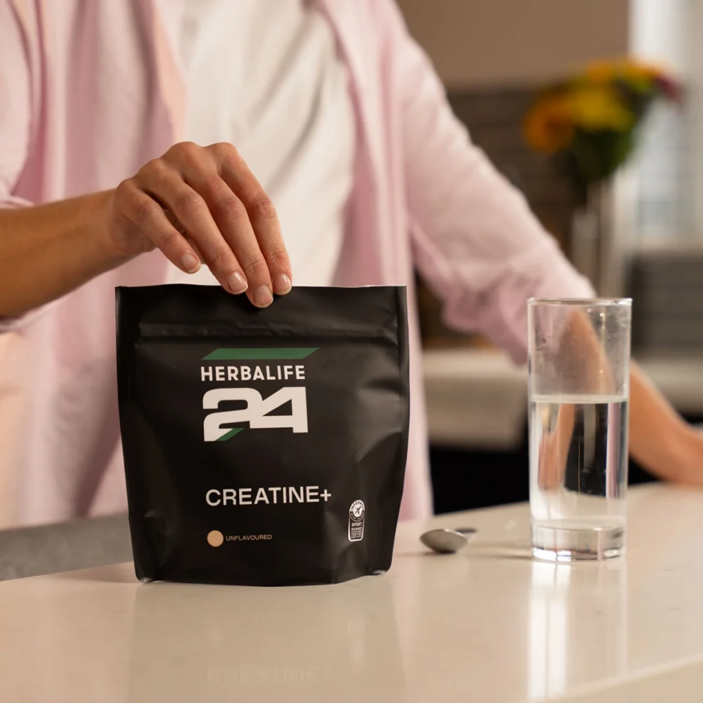 Herbalife24 Creatine+ unflavoured Pulverbeutel auf einer weißen Küchenarbeitsplatte neben einem Glas Wasser.